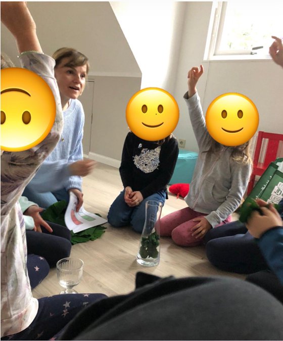 Nathalie Barth sitzt mit Kindern im Kreis auf dem Boden, ein Kind meldet sich, die Gesichter der Kinder sind verdeckt